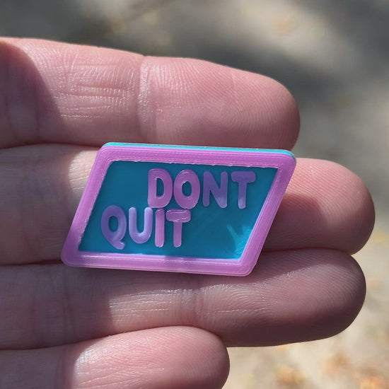 This is a short video that shows the transition of this pin when exposed to direct sunlight. The pin is a light blue hexagon with the words DONT QUIT in light purple. There is an opaque white border and letters that when put in direct sunlight reveal the message DO IT. 
