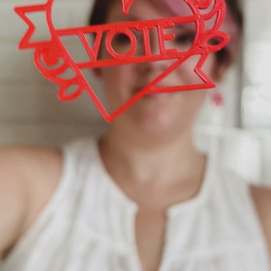 This is a video of Rebekah Thornhill, owner of R+D, showing a 3D printed earring. First she shows the earring close up. It is a bright red color and shaped as a heart with a banner twisting around it with roses on the sides. Writted on the banner is VOTE. After showing the earring, she puts it on and shows the size of the earrings when being worn. 