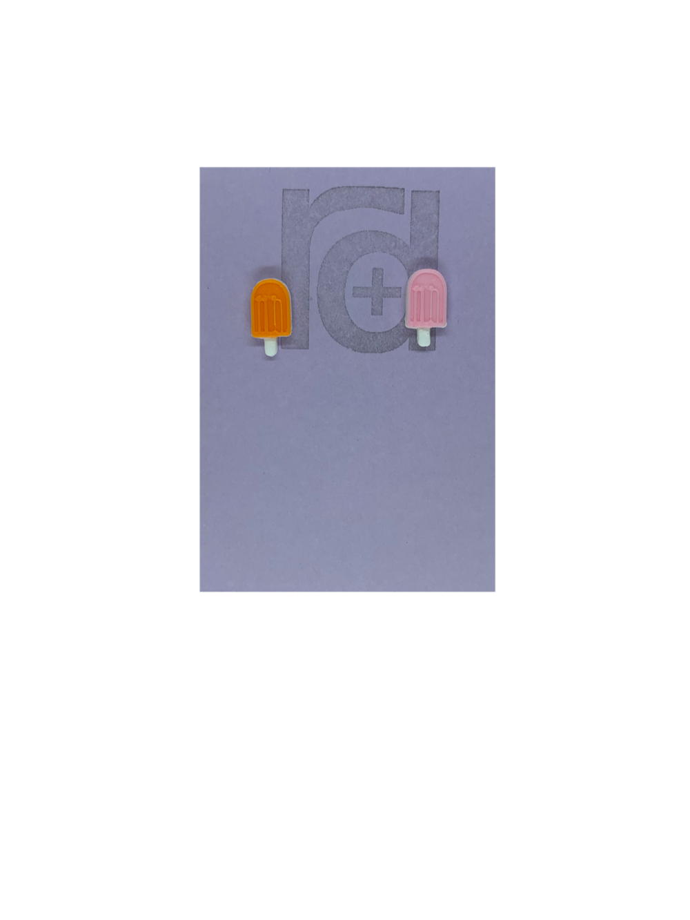 Two small and detailed R+D earrings are shown on a purple earring card. They are shaped as popsicles with white sticks. The earrings are mismatched, one popsicle is a vibrant orange and one is light pink