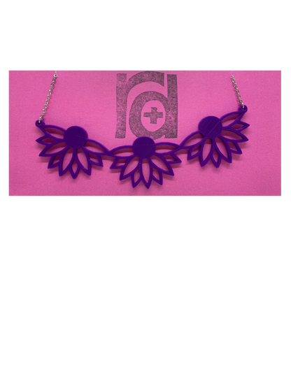 On a bright pink R+D necklace card is a 3D printed necklace. It is a bib necklace that stretches to have three daisys with petals stretching downward. This necklace is printed in a sustainable purple filament. 
