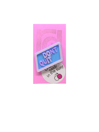 Shown on a pinkish lilac R+D card is 3D printed pin. It is in a light blue hexagon shape with purple writing that says DONT QUIT. There is also a opaque white layer on the pin. That layer is UV reactive and makes a border and reveals a hidden message in direct sunlight. Here it is shown out of the sunlight. 
