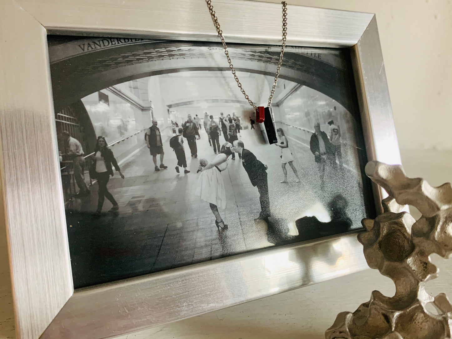 Hanging over a photo of two people bent and kissing in grand central station is a necklace with two 3D Printed pendants. The pendants are merlot red and black. When turned to the side, the pendant reveals that there is a heart on the dark red pendant and the date 9/20 on the black pendant. These pendants can be customized with different names or dates. 