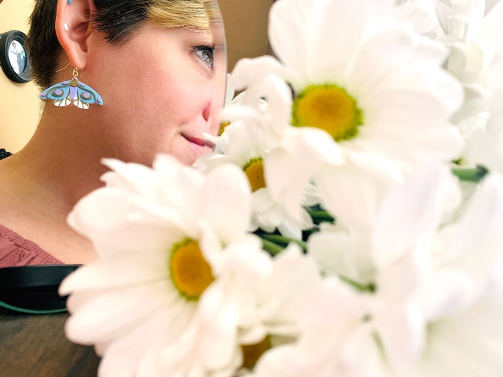 In the foreground is a bouquet of daisy flowers out of focus.  Further away, but in focus, is a mirror reflecting a person wearing a 3D Printed earring from R+D. The earring is a moth that has white lower wings, light blue larger wings, a light purple body and gold details on the wings and body. 