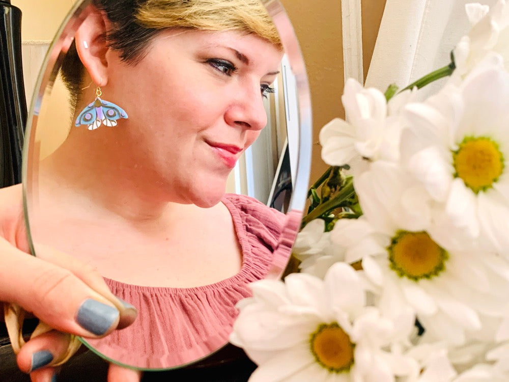 In the foreground is a bouquet of daisy flowers out of focus. Further away, but in focus, is a mirror being held up and reflecting a person wearing a 3D Printed earring from R+D. The earring is a moth that has white lower wings, light blue larger wings, a light purple body and gold details on the wings and body.