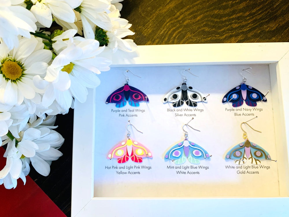 Shown in a white shadow box are six different colors of R+D 3D printed moth earrings. They are each held in place with pins like a shadow box in a natural history museum. The six colors are labeled: Purple and Teal Wings, Pink Accents; Black and White Wings, Silver Accents; Purple and Navy Wings, Blue Accents; Hot Pink and Light Pink Wings, Yellow Accents; Mint and Light Blue Wings, White Accents; and White and Light Blue Wings, Gold Accents.