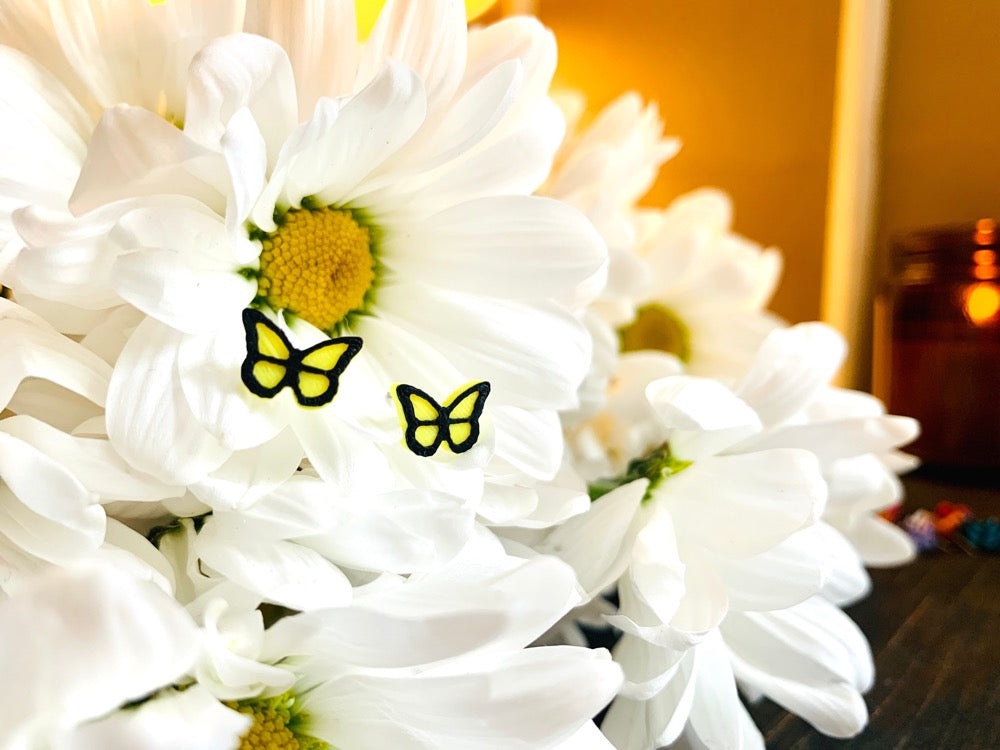 Resting in a bouquet of daisy flowers is a pair of R+D 3D Printed earrings. They are bright yellow butterflies with a black outline. These studs are small and delicate.