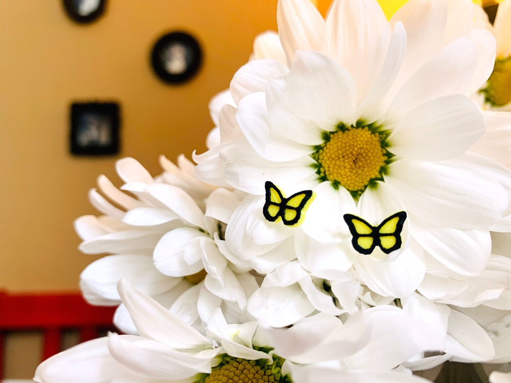 Resting in a bouquet of daisy flowers is a pair of R+D 3D Printed earrings. They are bright yellow butterflies with a black outline. These studs are small and delicate. 