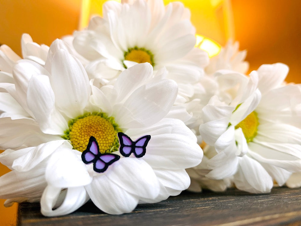 Resting in a bouquet of daisy flowers is a pair of R+D 3D Printed earrings. They are purple lavender butterflies with a black outline. These studs are small and delicate.