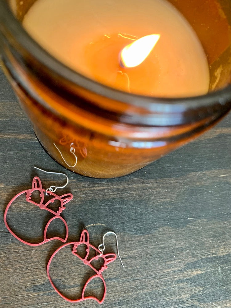 Laying next to a lit candle in a brown glass are two 3D printed R+D earrings. They are merlot red foxes that have big bushy tails curling over their bodies.