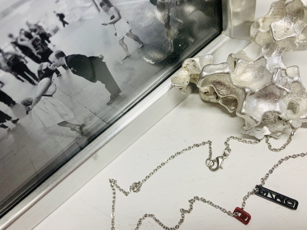 Sitting next to a photo of two people bent and kissing in grand central station is a necklace with two 3D Printed pendants. The pendants are merlot red and black. When turned to the side, the pendant reveals that there is a heart on the dark red pendant and the date 9/20 on the black pendant. These pendants can be customized with different names or dates. Edit alt text