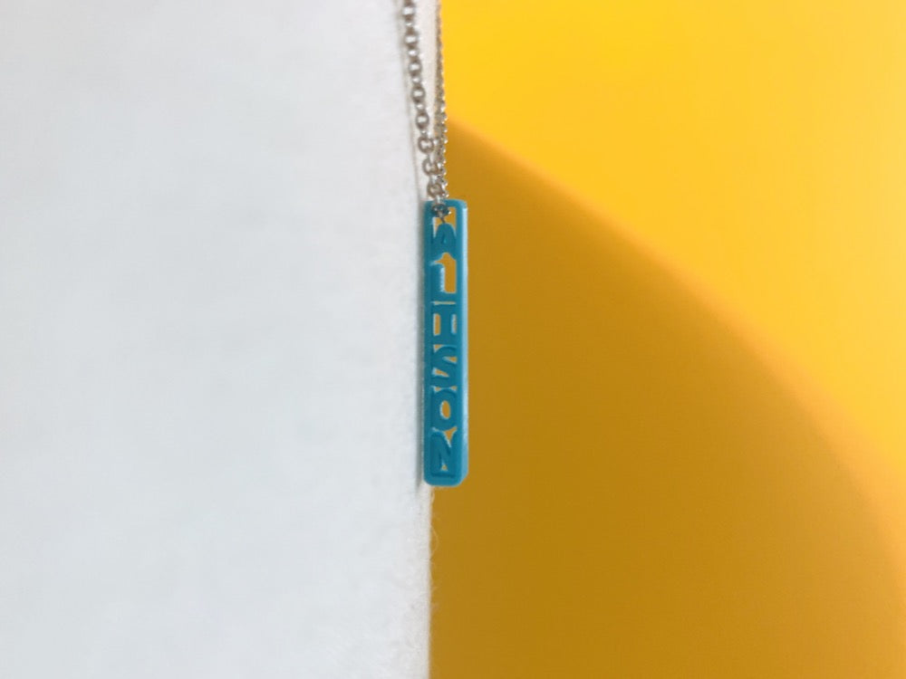 Hanging between a white background and a yellow background is a 3D printed pendant. It is a thin and tiny pendant that has the name ALISON printed on it in a bright teal plant based filament.