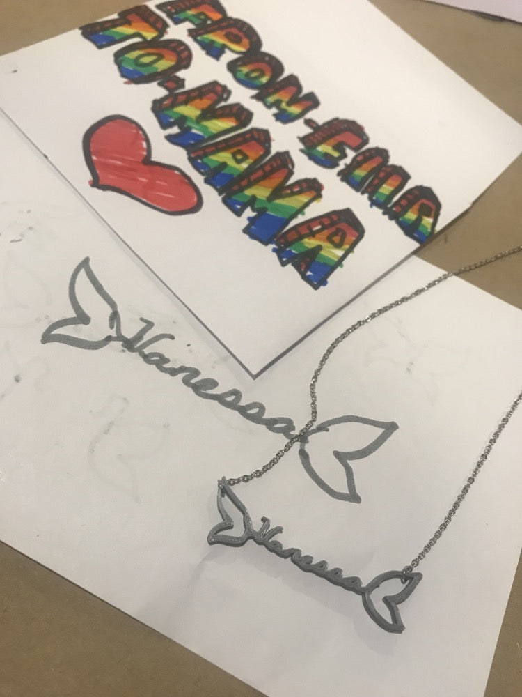 In the background is a hand drawn card. There's a drawing of a name and extra details that matches a 3D printed necklace that sits next to it.