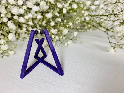 The background is filled with baby's breath flowers. In the foreground there are two 3D printed earrings. They are printed in a plant based purple filament and are shaped as triangle hoops. In the photo they are standing up to create a unique geometric structure. 
