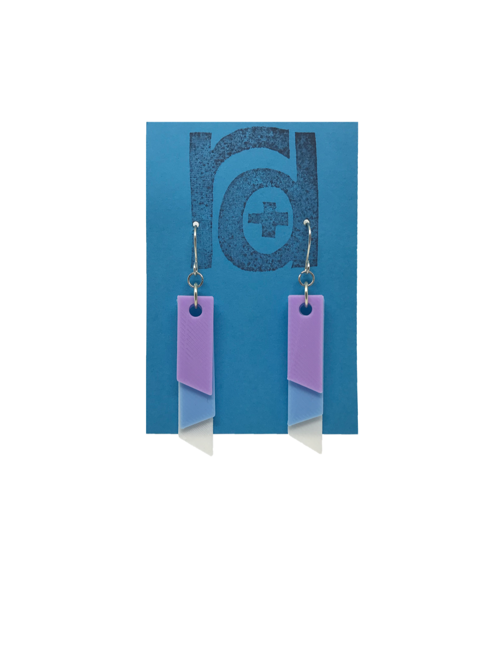 On a bright blue R+D earring card is a set of 3D printed earrings. The earrings are 3D printed with a plant based filament to make them sustainable as well as affordable. There are three banners hanging down; one is white, one is light blue, and the final is light purple. They are each different lengths so that the colors show.