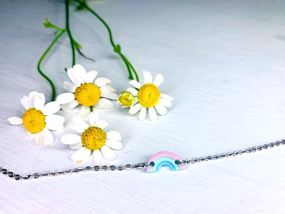 On a white background is a bracelet laid out in front of white and yellow chamomile flowers. The bracelet features a charm in the middle that is a small 3D printed rainbow made from pastel pink, mint green, and baby blue.