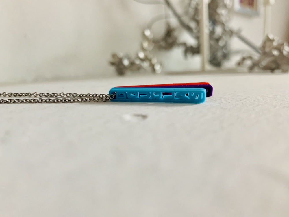 On a white background are three 3D printed pendants. They are each different colors and shaped as long rectangles with names in the center. When worn, they simply look like long hanging rectangles, but from the side, all the names are visible. This one has a teal pendant, a purple pendant, and a red pendant, but from this perspective the teal pendant is most visible. It reads Douglas.
