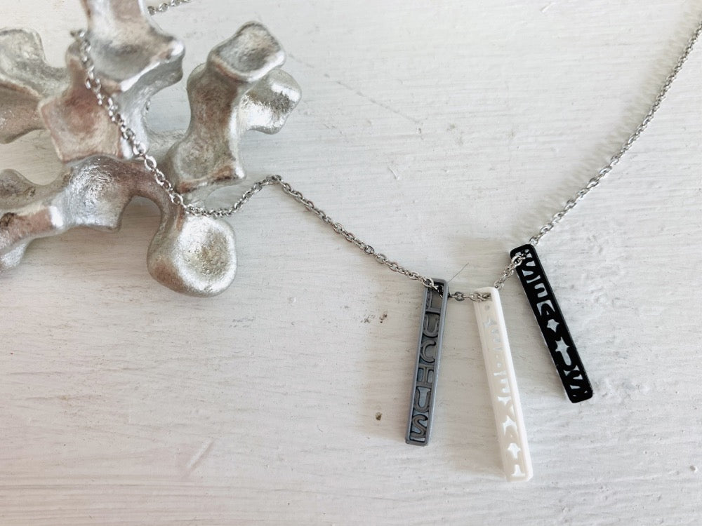 Shown on a white background with an aluminum sculpture on the side. There is a necklace with 3 3D printed pendants. The pendants are long and thin, in silver, white, and black. These are turned to the side to reveal three names: SEAMUS, LUCIUS, and REBEKAH. They can be customized to any name or word.