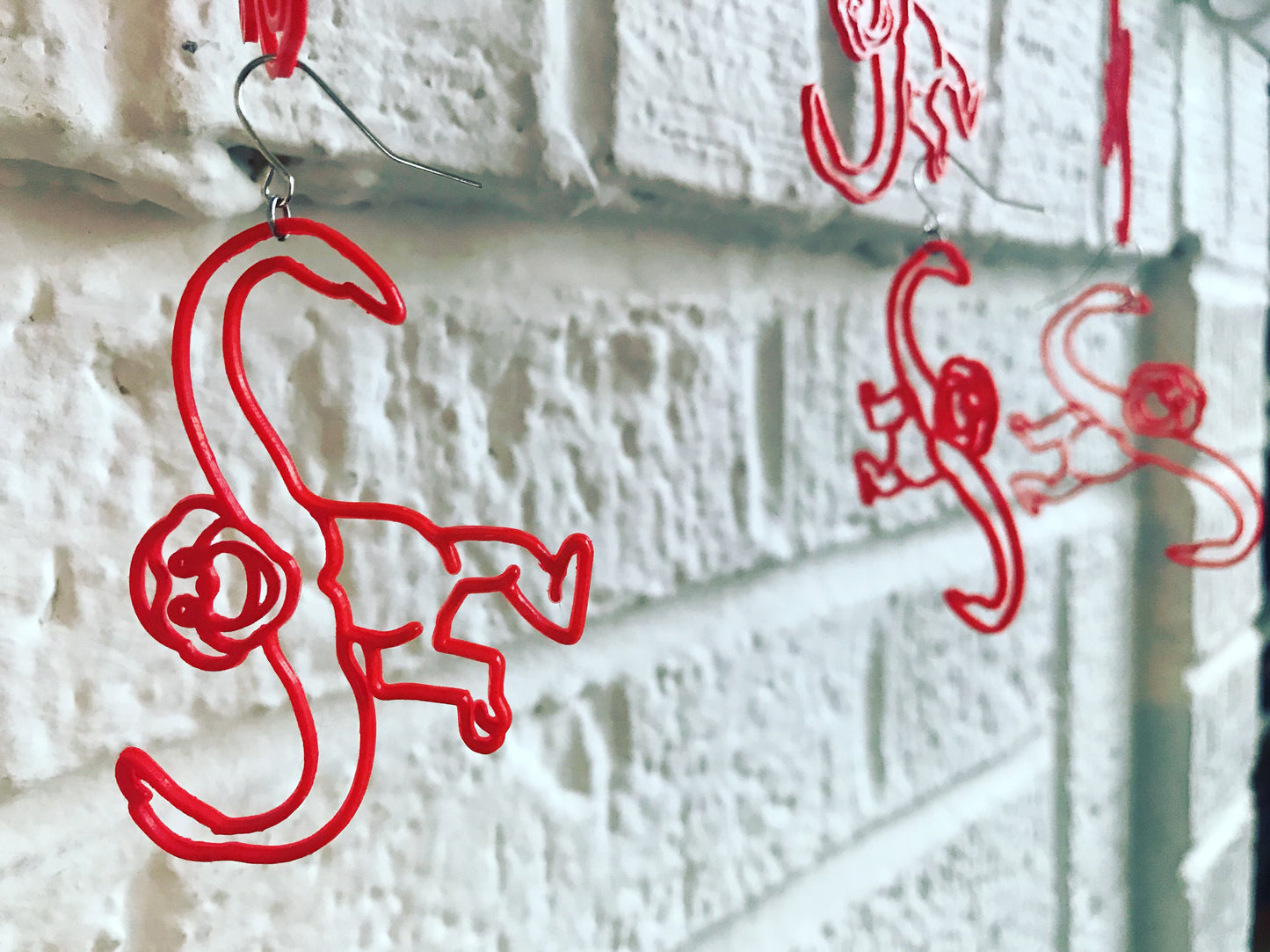 In the foreground is a bright red monkey earring. It is shaped like the classic barrel of monkeys toy where the monkeys have long twisting arms, a big smile, and a body that is horizontal when it is hanging so it looks like the monkey is actively swinging through the trees. In front of a white brick background, you can see other monkeys that are hanging down off of each other as though someone were playing the original game with these earrings.