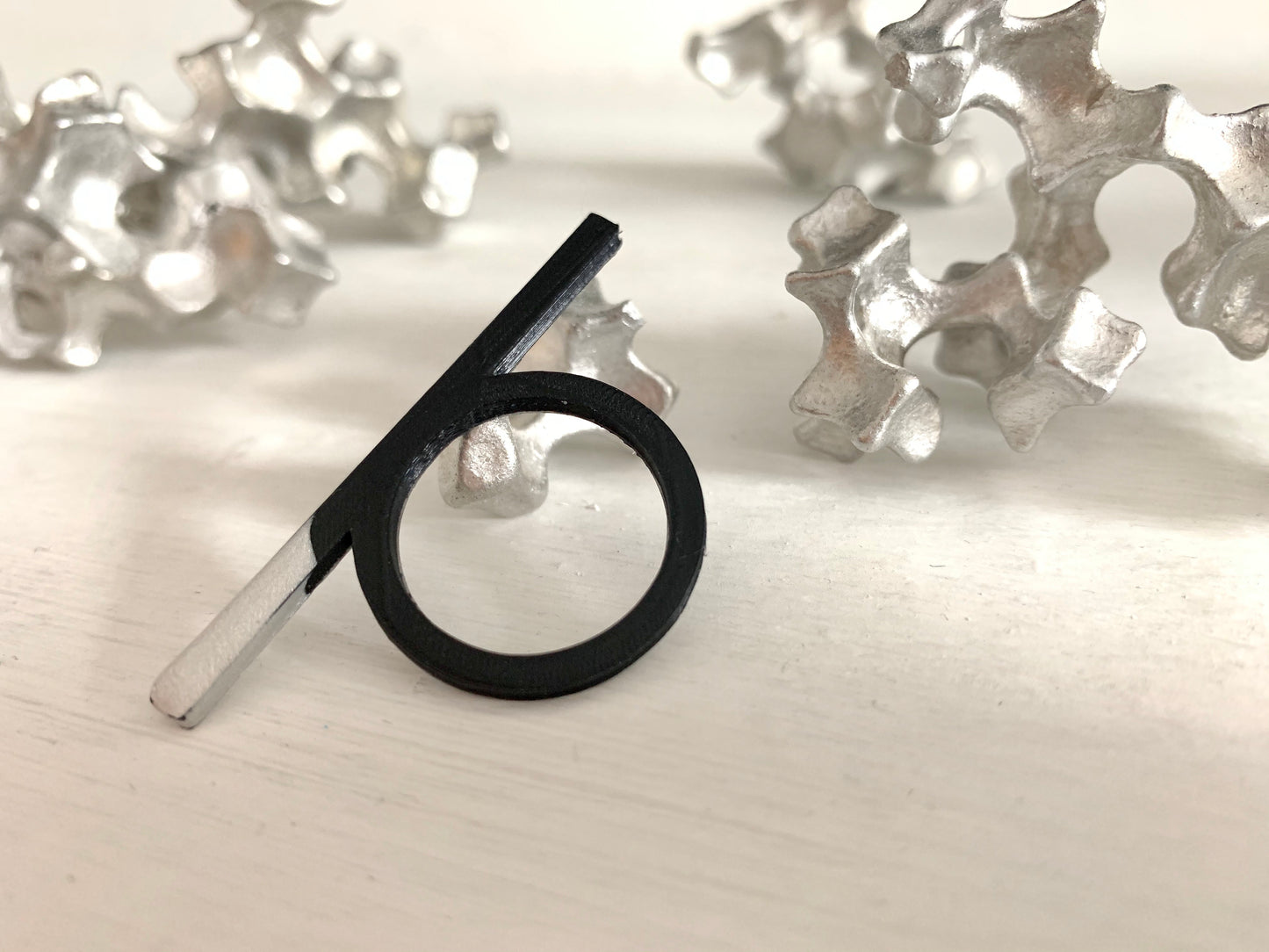 In the foreground is a black 3D printed ring that has a bar that extends over other fingers when being worn. It is dipped in a metallic silver paint on one side of that bar. In the background are cast aluminum sculptures that look like coral.