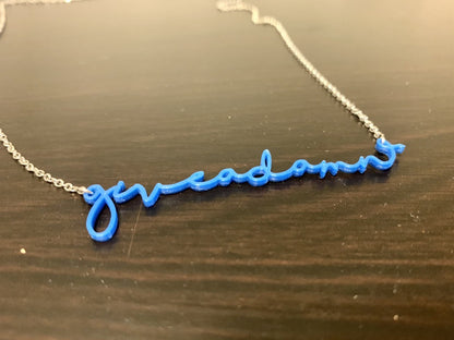 On a dark wood surface is a 3D printed pendant. In a modern cursive script it says give a damn  in a classic cobalt blue color.