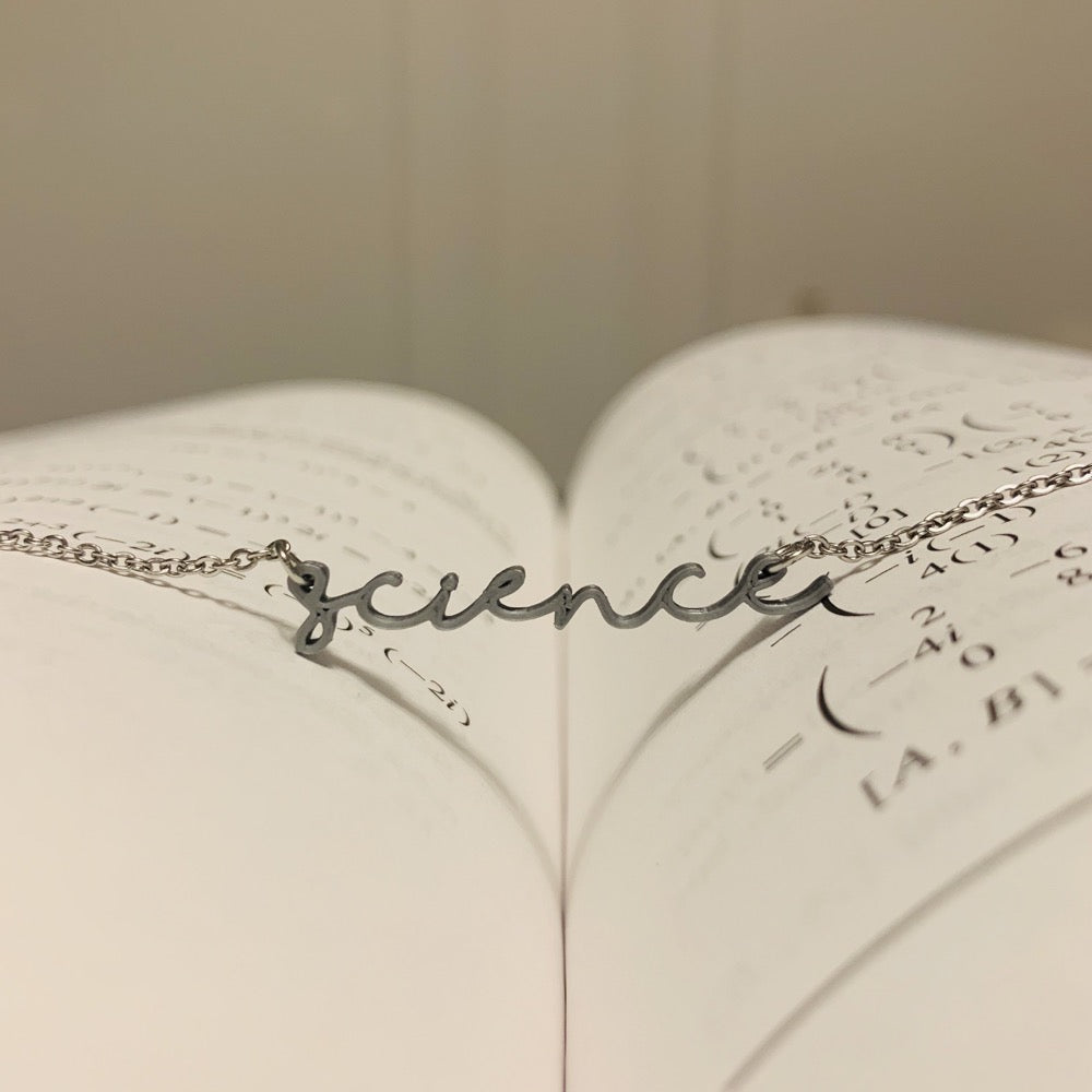 In the middle of an open book is a 3D printed pendant. In a modern cursive script it says science in a silvery grey color. 
