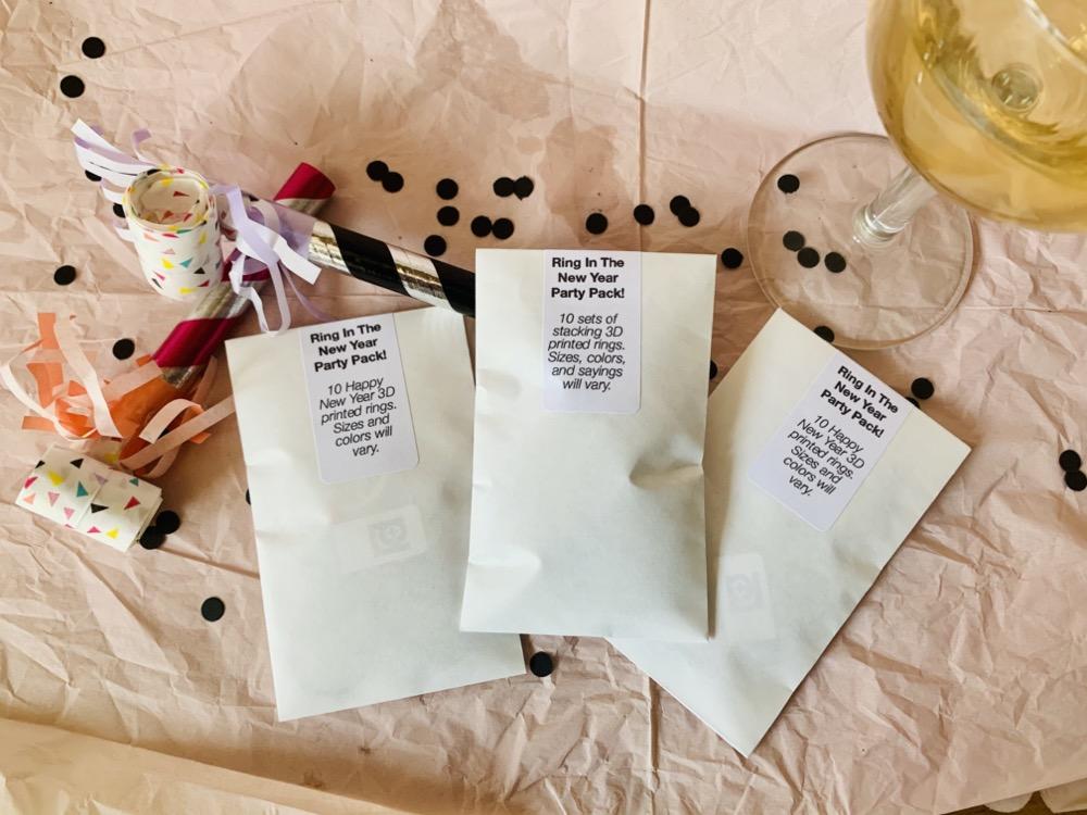 Laying across a light pink paper tablecloth is a party scene -- there is black confetti, two colorful noise makers, and a glass of champagne. Laying on top are three R+D party packs. These are shown closed and are white envelopes with a label across the opening. The label tells you that there are 10 New Year's Eve rings inside of varying colors and sizes.