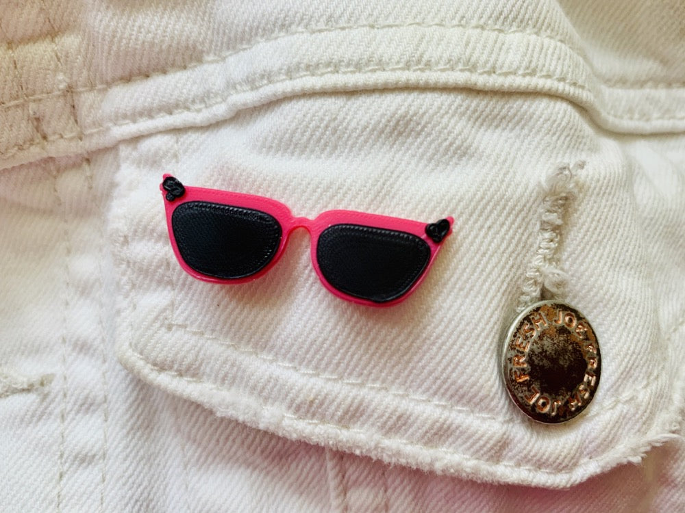 There is a close up of a white demin jacket pocket. An R+D pin is attached that looks like a pair of cat eye sunglasses. They are hot pink with black frames and black accents in the corners.