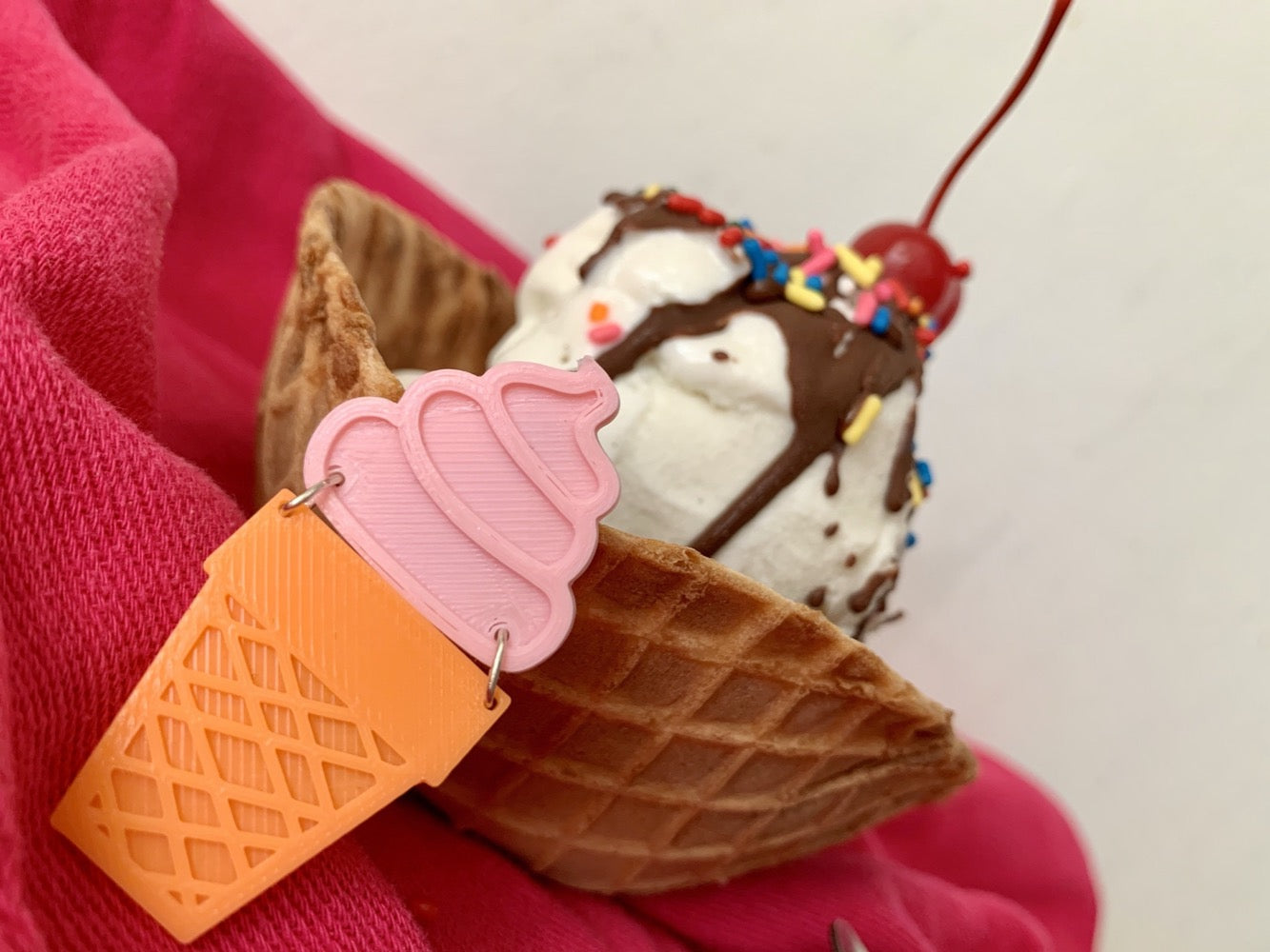 In the foreground is a sustainable ice cream pin that was 3D printed. The top of it is pink and looks like soft serve ice cream swirled into a peak. Below is an orange cone that looks like a classic sugar cone. In the background there is a waffle cone bowl holding an ice cream sundae with vanilla ice cream, rainbow sprinkles, chocolate sauce and topped with a bright red cherry.