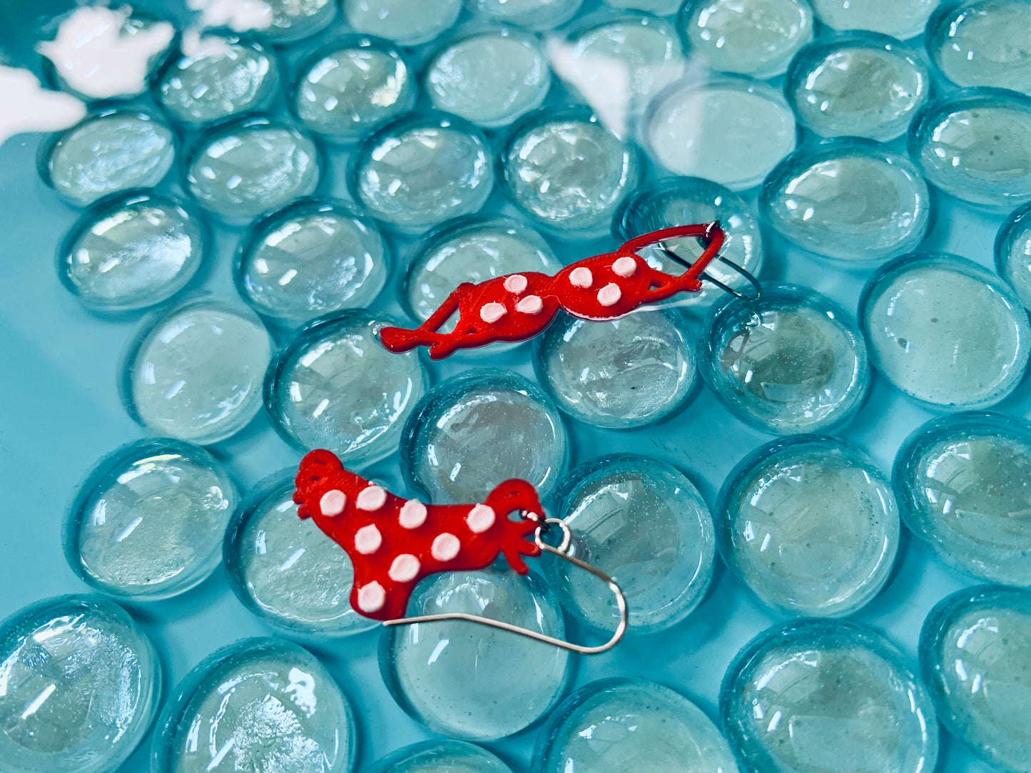 Floating in bright blue pool water are two 3D printed earrings. The earrings are shaped like a red bikini, one as a top, and one as a bottom. They have white polka dots covering them.
