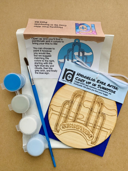 Shown laying on craft paper is the packaging and contents of this DIY Paint Kit. There is a paper envelope with instructions, a paintbrush, a set of 4 paint wells and a wall hanging with a laser cut NYC landmark.