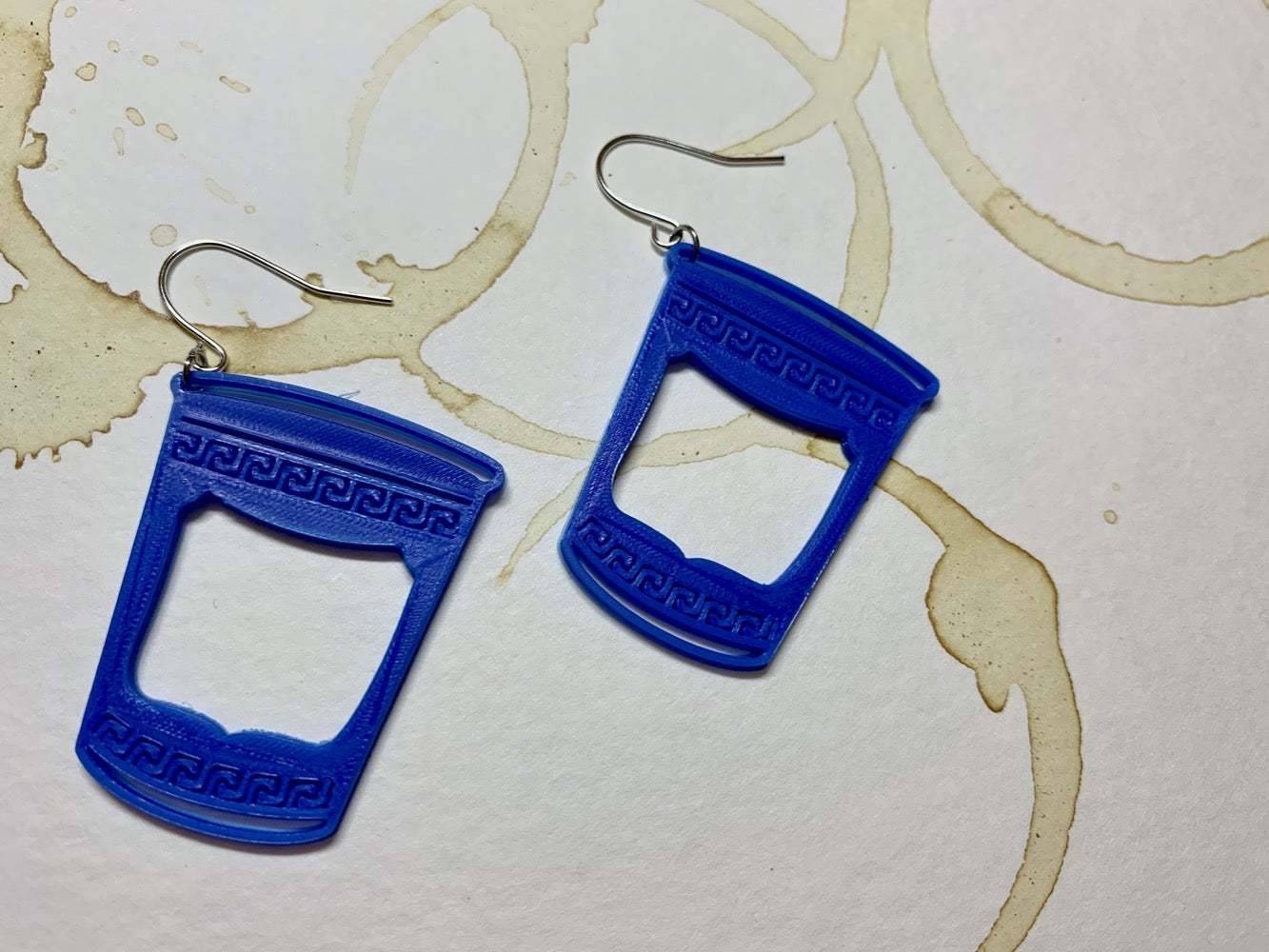 Two earrings that are made of a plant based plastic. They are in the shape of to-go coffee cups: The iconic ones from Greek diners or NYC bodegas with swirling borders and a big panel in the center. These earrings are resting on a paper that is stained with coffee rings overlapping one another.
