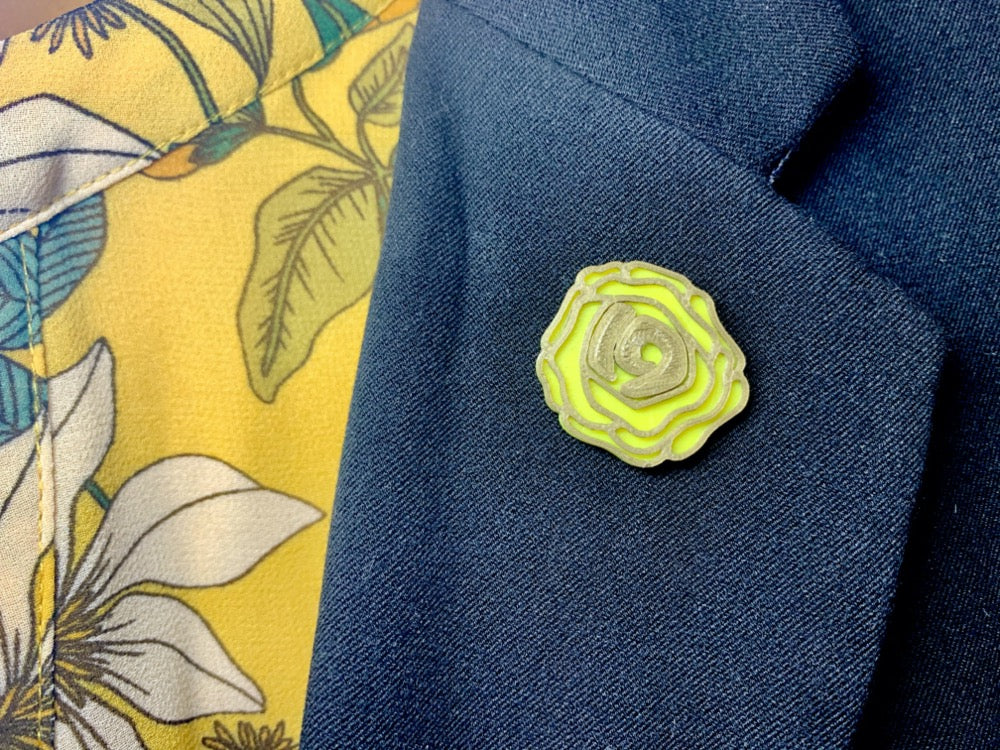 On a black suit lapel is a yellow rose pin. It's petals are outlined in gold and the center has the number 19. Those who were in support of the 19th amendment being ratified would wear a yellow rose.
