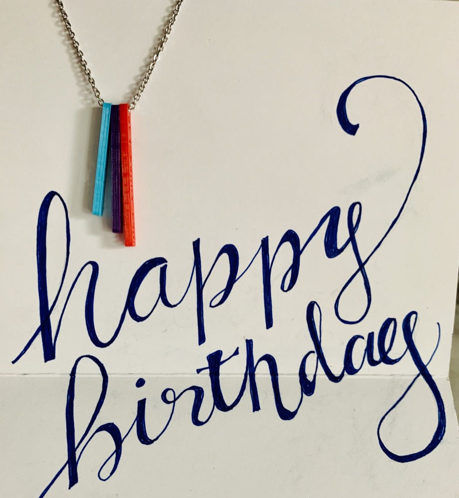 Hanging in a card with the words, happy birthday scrawled, is a necklace with three pendants. They are long rectangles in teal, purple, and red. When turned to the side, each pendant reveals a name. 