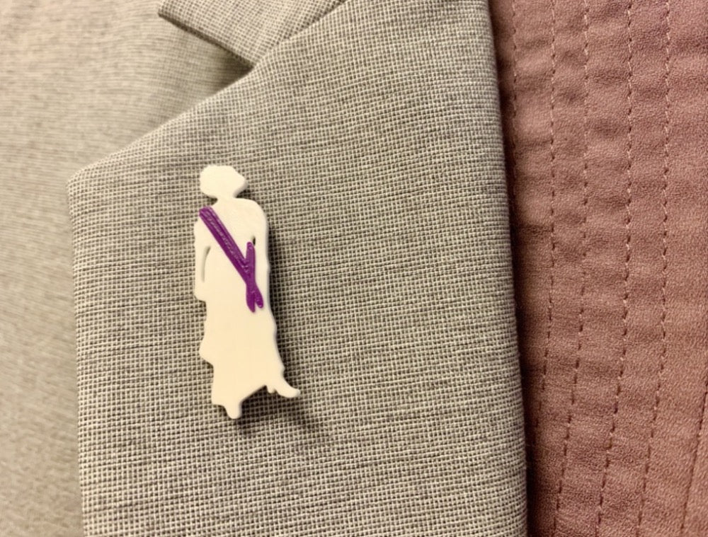 On the lapel of a grey suit is a 3D printed R+D pin. It is the silhouette of a woman from the 1910s wearing a long dress, hat, and heels. Her silhouette is in white, but there is a bright purple sash across her body to show she is a suffragette. 