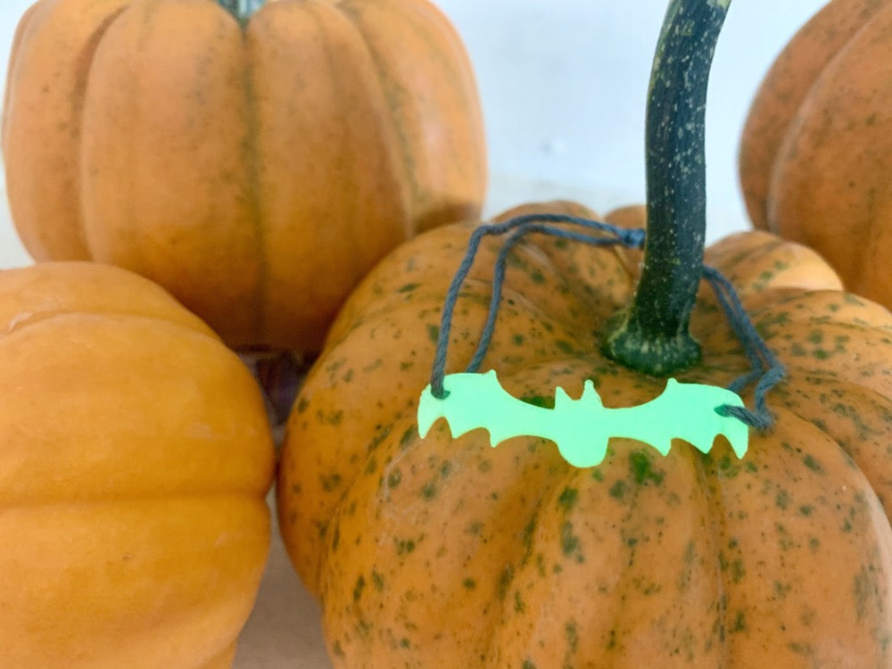 In this picture there are multiple pumpkins all around. On top of one is a bracelet. It has a dark grey adjustable cord that is holding on to a 3D printed bat that glows in the dark.