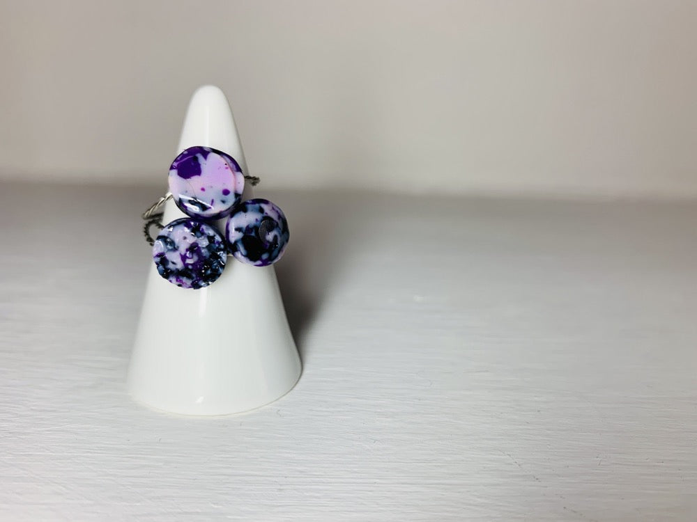 On a bright white background there is a white ring holder with three cast rings. The rings are all circle shapes, one smooth, one faceted, one bumpy like a geode. They are cast from recycled 3D prints in hues of purple black and white. It has the effect of marble or granite that is speckled. 