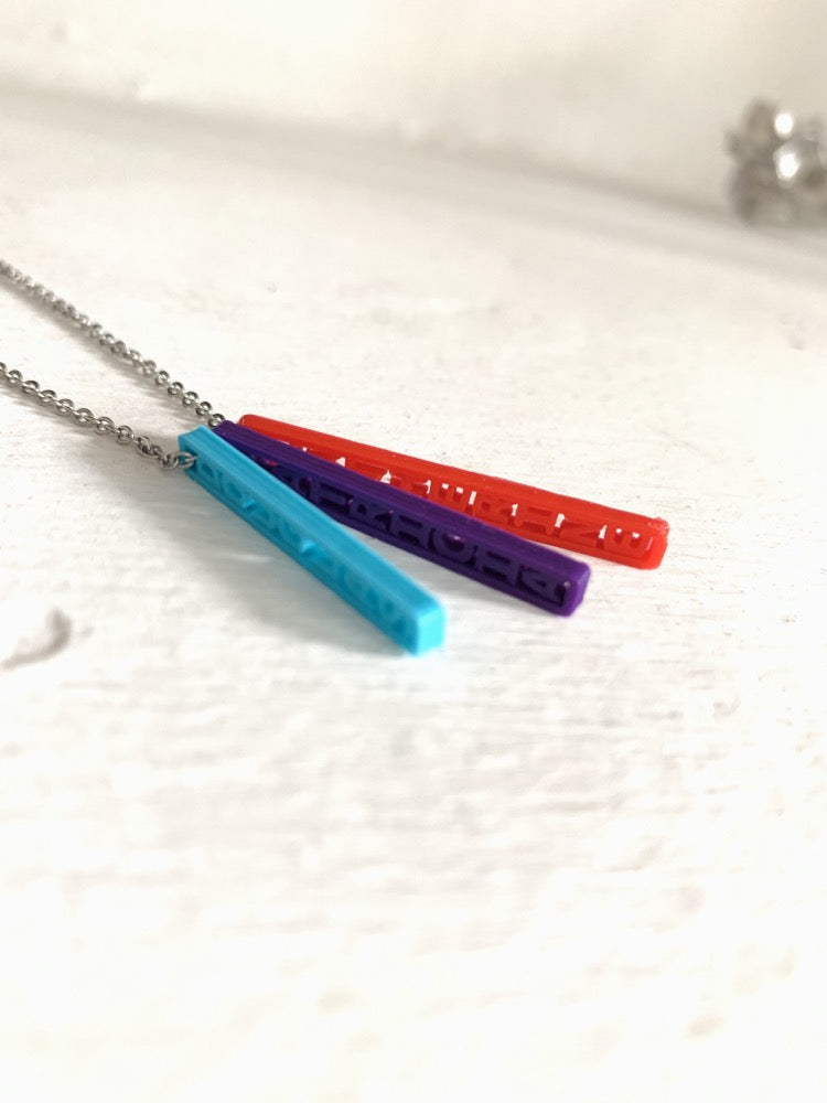 On a white background are three 3D printed pendants. They are each different colors and shaped as long rectangles with names in the center. When worn, they simply look like long hanging rectangles, but from the side, all the names are visible. This one has a teal pendant, a purple pendant, and a red pendant. 