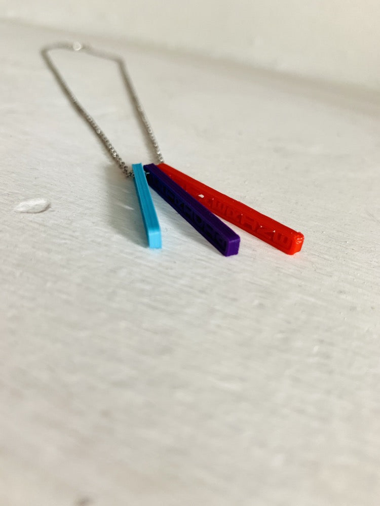 On a white background are three 3D printed pendants. They are each different colors and shaped as long rectangles with names in the center. When worn, they simply look like long hanging rectangles, but from the side, all the names are visible. This one has a teal pendant, a purple pendant, and a red pendant.