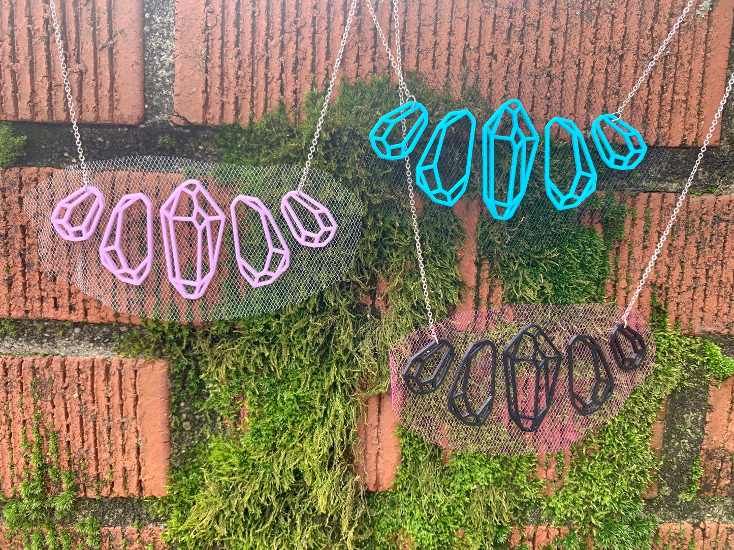 On a brick background with bright green moss are three necklaces. They are different colors of the same necklace. The R+D necklace features 5 crystal shapes; the center gem is the largestand they get smaller as they move to the sides symmetrically. The gems don't touch, but are connected by tulle fabric that is embedded in the 3D print. The necklaces are pictured with white tulle and light purple filament, black tulle and teal filament, and hot pink tulle and black fillament.