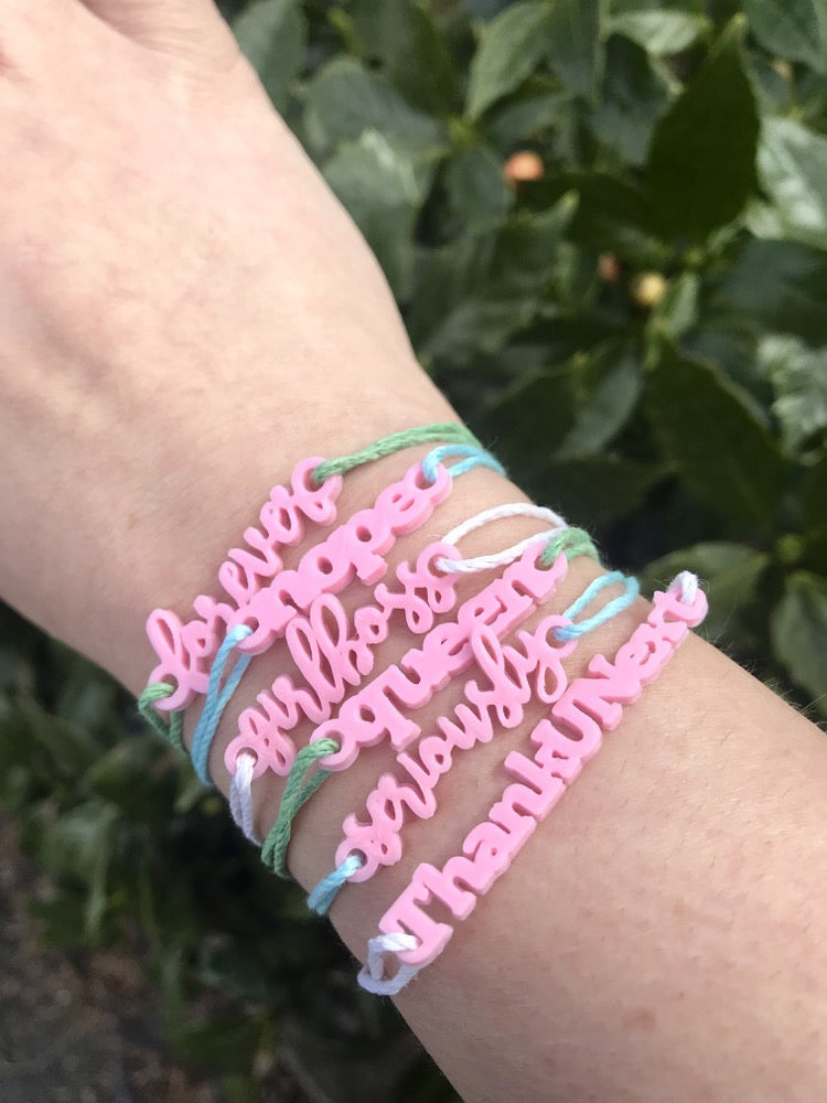 In the foreground are six light pink 3D printed bracelets. From top to bottom thy say forever in cursive, nope in block letters, girl boss in cursive, queen in block letters, seriously in cursive, and ThankUNext in block letters.