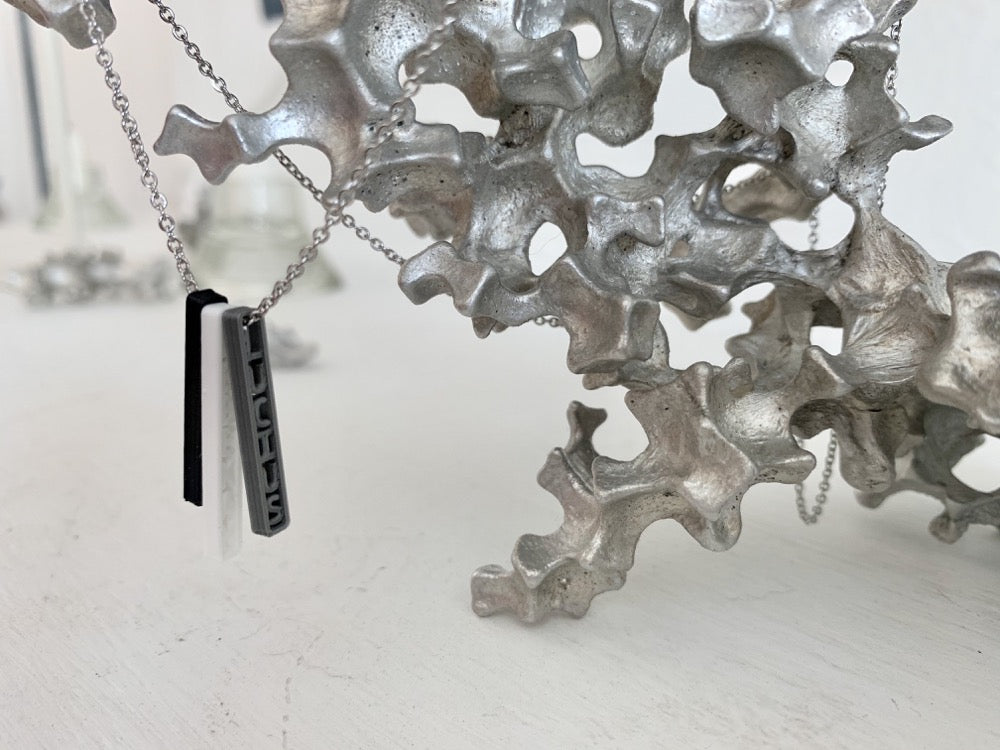 Shown on a white background with an aluminum sculpture on the side. There is a necklace with 3 3D printed pendants. The pendants are long and thin, in silver, white, and black. When turned to the side, names are visible on the pendants. One the silver pendant, the name LUCIUS is visible.