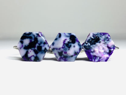 On a bright white background are three rings. They are all cast using recycled 3D prints. They have a speckled or granite like appearance with shades of white, black, purple, and light purple plant based filaments. They are hexagons in shape; one has a smooth surface, another a faceted surface, and finally a bumpy geode surface. 