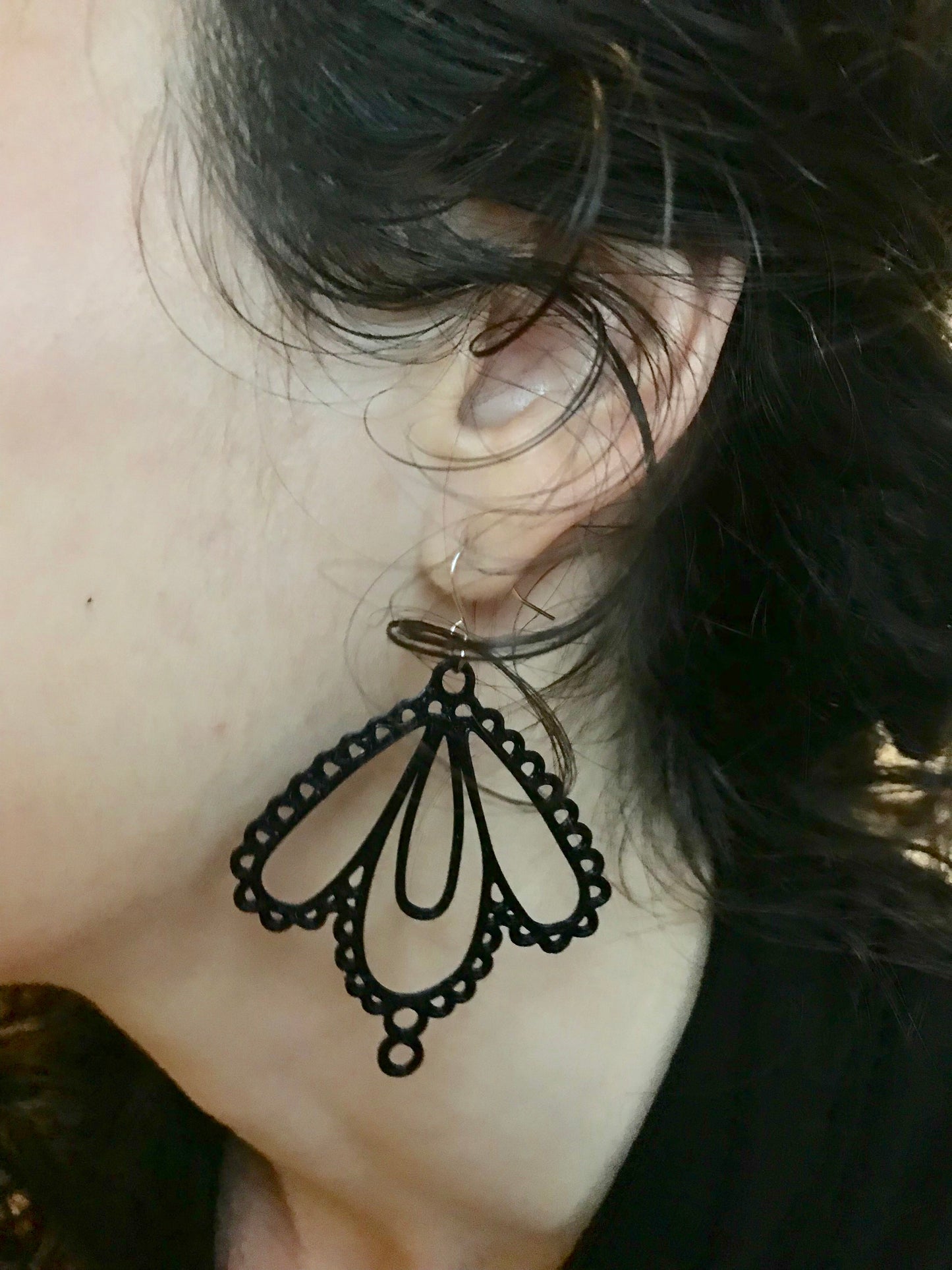 A close up of a woman with curly hair's ear and the black earring she is modeling. The earring has multiple tear drop shapes that are accented with more circles and loops to look like lace.