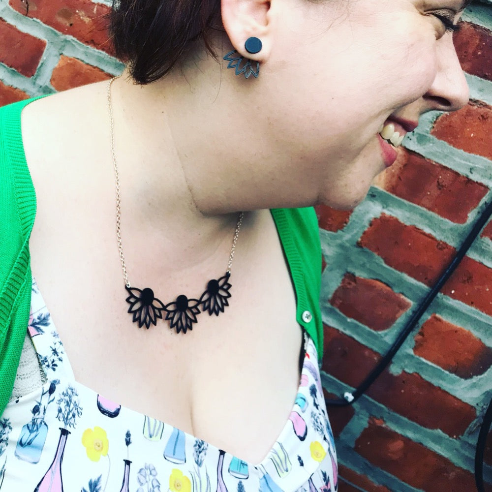 A woman is shown looking to the side while wearing a 3D printed necklace. The necklace is a bib necklace that stretches to have three daisys with petals stretching downward. This necklace is printed in a sustainable black filament.