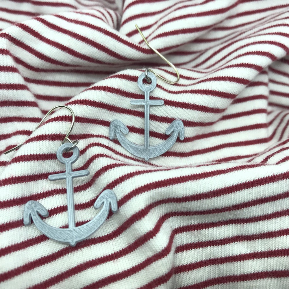 Two silver earrings are shown on red and while striped fabric. The earrings are shaped like boat anchors and 3D printed.