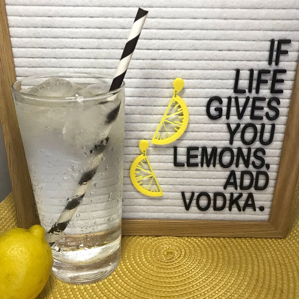 There is a white felt letterboard on a yellow placemat with a lemon and a tall glass of ice, tonic, and vodka. There is a black and white paper straw in the glass. The felt board says, If life gives you lemons, add vodka. It has two R+D earrings hanging off the board that are bright yellow and shaped like citrus slices and drops of juice.