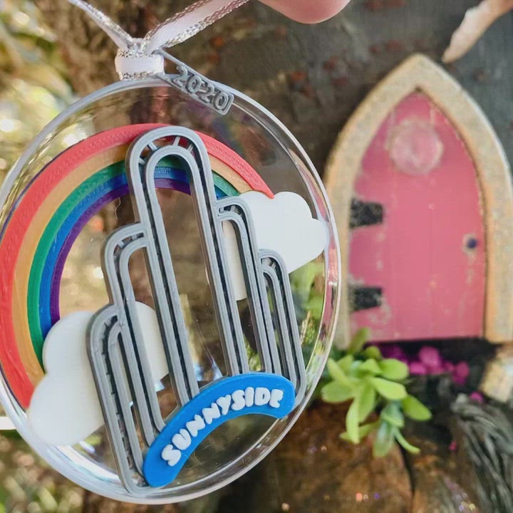 Outside of a pink fairy door in a tree, a hand dangles our limited edition ornament for 2020--It features the art deco arch in Sunnyside, Queens, NYC, with a rainbow and clouds behind it. In the video the ornament is turned, revealing a small fairy door at the bottom of the arch. 
