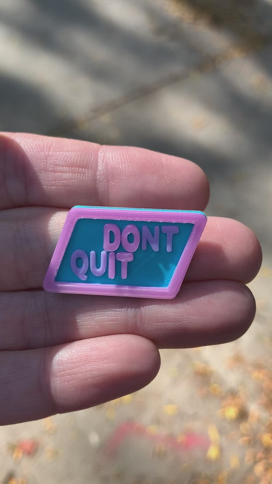 This is a short video that shows the transition of this pin when exposed to direct sunlight. The pin is a light blue hexagon with the words DONT QUIT in light purple. There is an opaque white border and letters that when put in direct sunlight reveal the message DO IT. 