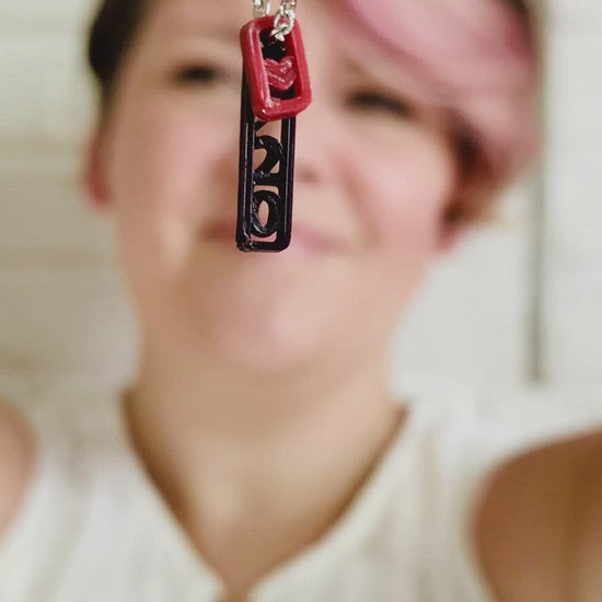This video starts with a 3D printed pendant necklace being held close to the camera. There is a dark red merlot pendant that has a heart and behind it is a black pendant, it reads 9/20 but only the 20 is visible from this viewpoint. The person holding the necklace pulls it away from the camera and some into focus as she puts it on and shows how the necklace looks when being worn. It is a very simple and delicate pendant. 