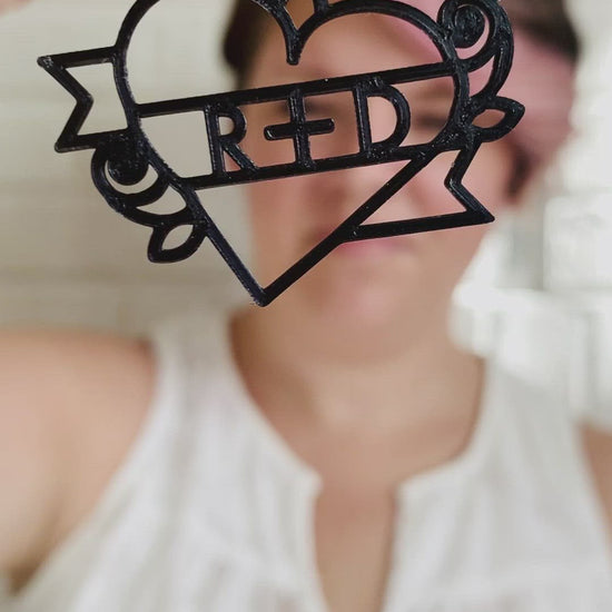 This is a video of Rebekah Thornhill, owner of R+D, showing a 3D printed earring. First she shows the earring close up. It is a bright red color and shaped as a heart with a banner twisting around it with roses on the sides. Writted on the banner is R+D. After showing the earring, she puts it on and shows the size of the earrings when being worn.